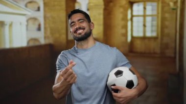 Üniversite koridorunda futbol topunu tutarken parmakları gülümseyerek kalp hareketi yapan genç bir adam.