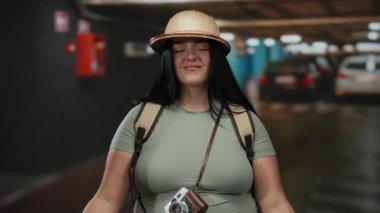 Kameralı ve sırt çantalı genç bir kadın kapalı otoparkta mutlu bir şekilde gülümsüyor..
