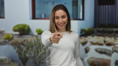 Açık bir günde, canlı bir sokak ortamını yansıtan bitkilerle dolu modern beyaz bir binanın önünde gülümseyen ve işaret eden bir kadın..