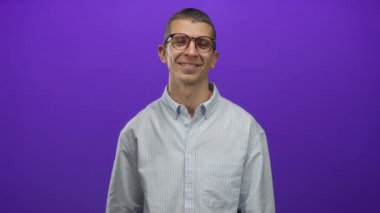 Gözlüklü, çizgili tişörtlü, parlak mor arka plan önünde taş gibi bir jest yapan adam, coşkulu tavır sergiliyor ve neşeli bir ifade sergiliyor, sade duvarda izole edilmiş..