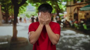 Kırmızı tişörtlü genç Çinli adam şehir caddesinde yapraklı ağaçlar ve arka planda bulanık insanlar görünce şaşırdı..