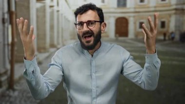 Sakallı İspanyol bir adam, eski bir üniversite kampüsünde şaşkın bir ifade sergiliyor, tarihi mimari ile çevrili, şaşkınlık ve hayal kırıklığı hissi uyandırıyor..