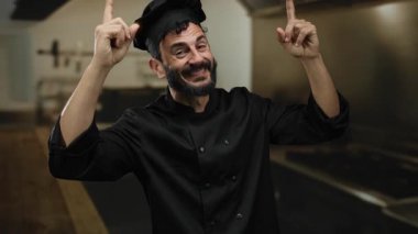 Profesyonel bir mutfağa işaret eden sakallı İspanyol erkek şef geleneksel siyah şef üniforması giyerken coşku ve uzmanlık gösteriyor..