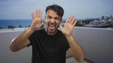 Genç İspanyol adam açık havada komik suratlar yapıyor ve arka planda okyanus manzarası olan bir terasta gülüyor. Açık gökyüzünün altında neşe ve neşe taşıyor..