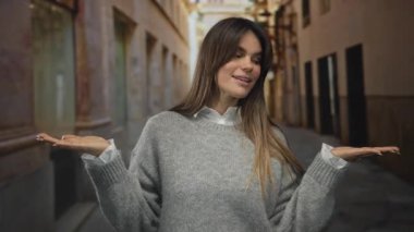 Dar bir caddede kolları açık gülümseyen genç İspanyol kadın gri kazak giyiyor.