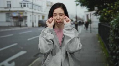 Bir kadın sokakta dikiliyor, neşeli bir ağlama hareketi sergiliyor, günlük bir kapüşonlu giyiyor, şehir merkezinde bulanık bir arka planda duyguları tasvir ediyor..