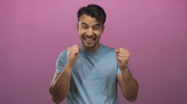 Pembe arka planda kapalı yumruklarla heyecanı ifade eden genç bir adam, mavi bir gömlek giyiyor ve neşe ve coşku gösteriyor..