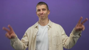 Mor duvara gülümseyen bir adam beyaz gömlekli ve bej ceketli el hareketleriyle neşeli bir ifade ve izole bir arka planda sıradan bir tarz sergiliyor..
