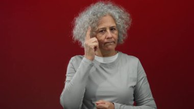 Göz alıcı kırmızı bir zemin üzerinde duran, gri saçlarını ve düşünceli ifadesini sergileyen, kavramsal kullanım için mükemmel bir şekilde izole edilmiş bir kadın..