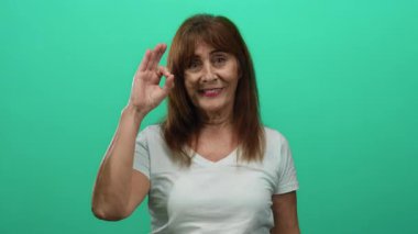 Kıdemli İspanyol kadın yeşil arka planda hoş bir jestle gülümsüyor izole bir ortamda günlük beyaz gömlek giyerek neşe ve pozitiflik sergiliyor.