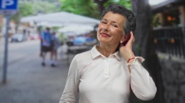 Gri saçlı yaşlı bir kadın, şehir sokaklarında elleri ile saçlarını tarıyor. Arka planı bulanık insanlar ve kafe masaları, bir şehirde mutluluk ve zarafet yayıyor.. 