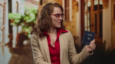 Alman pasaportlu bir kadın, güler yüzlü, kırmızı gözlüklü, bej ceketli, kimlik ve seyahat belgelerini ön plana çıkarıyor..