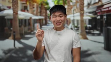 Beyaz gömlekli genç adam, güneşli bir şehir sokağında palmiye ağaçlarıyla eğlenceli bir jest yapıyor, açık hava kentsel manzarasını çekiyor..