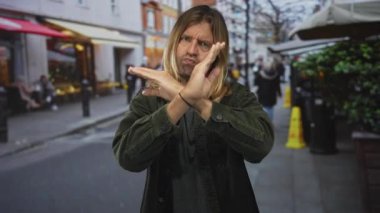 Uzun sarı saçlı, yeşil kadife ceketli bir adam kalabalık bir caddede x jestinde kollarını kavuşturuyor; inkar.