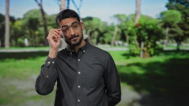 Parkta gözlüklü İspanyol bir adam, koyu renk bir gömlek giyiyor, güneşli bir günde güneşli ağaçların altında kendine güvenini ve tarzını gösteriyor..