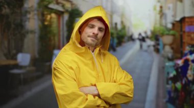 Şehrin arka planı bulanık, sarı yağmurluklu, çapraz kollu, kendine güvenen bir adam, açık havada şehir ortamında ciddi bir yüz ifadesi sergiliyor..