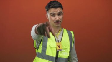 Yansıtıcı yelek giyen genç İspanyol adam izole edilmiş turuncu arka plana karşı gönüllü olarak işaret ediyor ve dost ifadesiyle alkışlıyor..