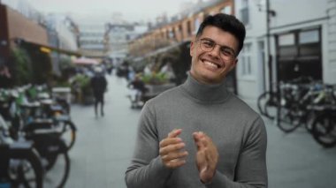 Canlı bir şehir caddesinde el çırpan genç bir adam gözlüklü ve arka planda insan ve bisikletli bir süveter giyen canlı bir şehir çevresi sergiliyor..