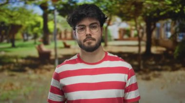 Ağaçlar, banklar ve açık hava manzarası ile çevrili güneşli yeşil bir parkta çizgili gömlek jestleri ile genç İspanyol adam, bir hoş geldin jesti önerir.