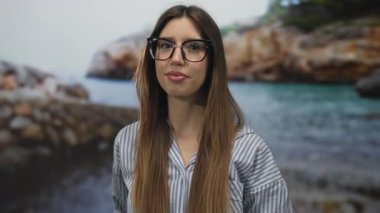 Çizgili gömlek giyen ve büyük gözlüklü bir kadın güneş ışığı altında kayalık sahil şeridini işaret ediyor..