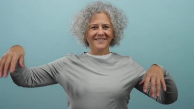 Neşeli mavi arka planda dans eden kadın enerjik ruhunu ve gri kıvırcık saçlarını sergiliyor..