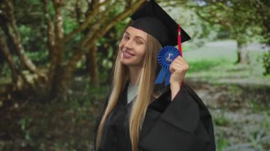 Mezuniyet elbisesi içinde elinde birinci sınıf bir kurdele, ağaçların arasından süzülen güneş ışığıyla yeşil bir parkta gülümseyen genç bir kadın..