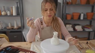 Kadın seramik vazoyu çömlek atölyesinde dönen çarkın üzerine kilden kaplanmış ellerle boyuyor; yaratıcılık odaklı sükunet.