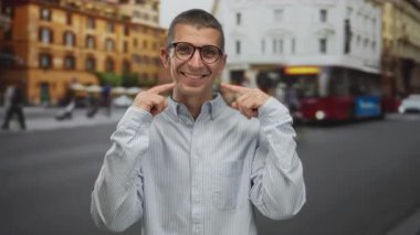 Kentsel caddede gözlüklü bir adam gülümsüyor. Şehrin arka planında parmaklarıyla gülüyor. Çevrede mutluluk ve neşe sergiliyor..