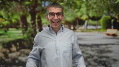 Güneşli bir parkta gülümseyen, gözlüklü ve çizgili tişörtlü, yemyeşil ağaçlar ve patikalarla çevrili, neşeli bir açık hava yaratan bir adam..