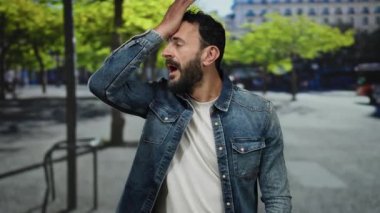 Bir şehir caddesinde duran İspanyol sakallı adam, şaşırmış ve düşünceli görünüyor, bir kot ceket ve canlı yeşil ağaçların altında beyaz gömlek giyiyor şehir arka planında.