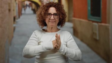 Şehrin caddesinde duran kıvırcık saçlı ve gözlüklü İspanyol kadın yumruklarıyla neşeli bir jest yapıyor. Açık hava kentsel ortamında mizah ve kendine güvenini ifade ediyor..