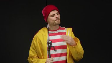 Sarı yağmurluklu genç adam siyah arka planda olta tutarken uzanan eline tuhaf bir ifadeyle bakıyor.