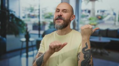 Sakallı ve dövmeli kel bir adam otel ortamında içeriyi işaret ediyor, rahat ve davetkar bir atmosferi taşıyor..