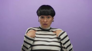 İzole edilmiş mor bir arka plana dikkat çeken bir kadın sürpriz yaparken çizgili bir süveter, kalın küpeler ve kısa saç stili giyiyor.