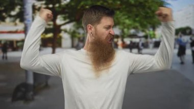 Genç sakallı adam arka planda bulanık yayalar ve kafelerle hareketli şehir caddesinde kollarını esnetiyor..