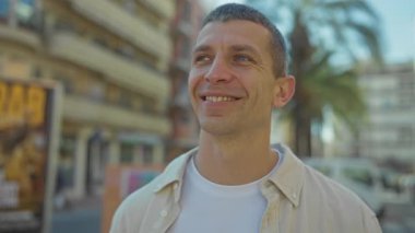Güneşli bir günde açık havada gülümseyen bir adam. Palmiye ağaçları, şehir binaları ve arka planda açık bir gökyüzü. Şehir ortamında mutluluğu taşıyor..