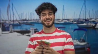 Limanda gülümseyen İspanyol bir adam arka planda bulanık tekne ve gemilerle İsviçre frangı tutarken sakin bir deniz manzarası yakalıyor..