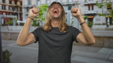 Uzun sarı saçlı bir adam şehir caddesinde her iki işaret parmağını yukarı kaldırırken güler yüzlü, iyimser..