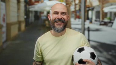Şehir sokağında elinde futbol topu tutan sakallı mutlu kel adam kamerayı işaret ediyor.