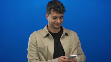 Bir stüdyo sahnesinde, mavi bir arkaplanda mesajlaşmak için akıllı telefon kullanan İspanyol bir genç adamla mesajlaşırken adam telefonu elinde tutuyor..