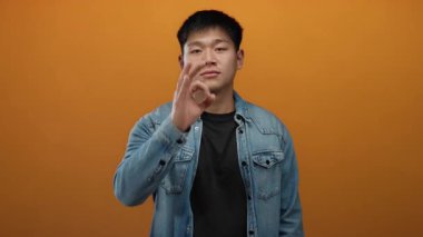 Kot ceket giyen genç adam izole edilmiş turuncu arka planda iyi bir jest yapıyor, özgüven ve iletişim gösteriyor..