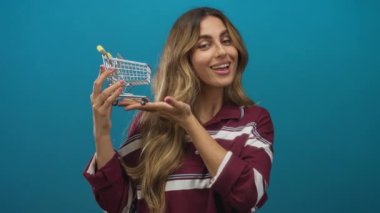 Mavi stüdyoda iki eli de minyatür alışveriş arabası tutan genç sarışın bir kadın var..