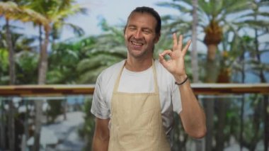 Bej önlük giyen gülümseyen sarışın adam lüks otel binasında el hareketi yapıyor. İyimserlik konukseverliğine hoş geldin..