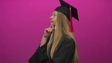 Mezuniyet şapkalı ve cüppeli bir kadın pembe arka planda yalnız düşünüyor.