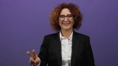 Gülümsemesi ve kendine güveni olan bir kadın canlı mor bir arka plana karşı barış işareti gösteriyor. Pozitif enerji ve iş kıyafetlerinde zarif bir tarz sergiliyor..