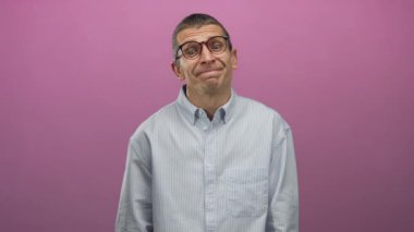 Gözlüklü, çizgili tişört giyen beyaz bir adam pembe arka plana karşı duruyor. Omuz silkme, izole ve gayri resmi bir ortam ile kafa karışıklığı gösteriyor..