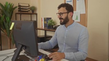 Sakallı İspanyol bir adam modern ofis ortamında eğleniyor. Masaüstü bilgisayarının önünde neşeyle gülüyor ve dans ediyor. Kapalı alanda canlı bir çalışma ortamı yaratıyor..