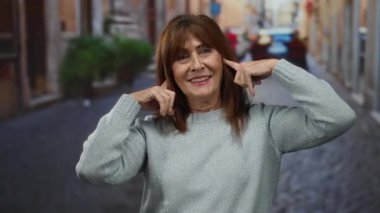 Yaşlı İspanyol kadın şehir sokaklarında kulaklarını tıkıyor, stres veya hayal kırıklığını dile getiriyor, gri bir kazak giyiyor, şehir hayatını ve duygularını yakalıyor..