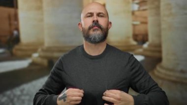 Sakallı ve dövmeli kel bir adam şehir mimarisi ile çevrili bir caddede güven içinde duruyor ve kırsal Avrupa sütunlarını gösteriyor..