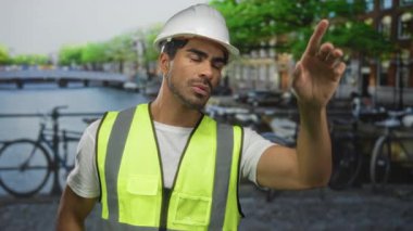 Sarı güvenlik yeleği ve beyaz şapkalı İspanyol adam inşaat halindeki binayı işaret ediyor..